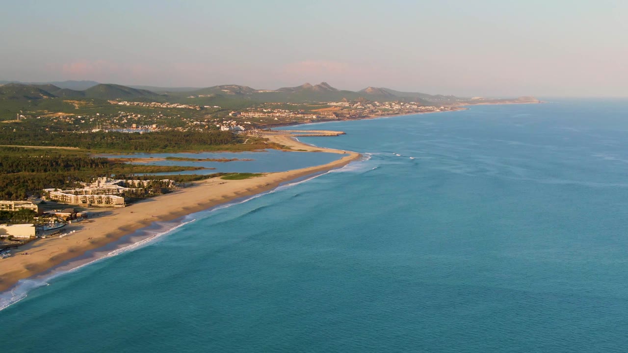 Wide San Jose del Cabo aerial frame with more open turquoise water to the right, a sandy shoreline and white surf to the left, small boats farther along the coast, low buildings, lagoons, hills, and no visible people.