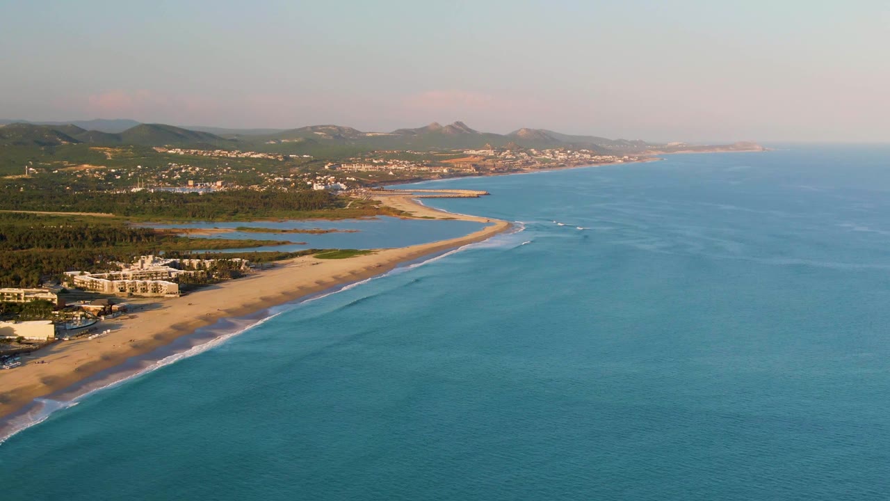 High aerial view of the San Jose del Cabo beach with the shoreline angling through the left side, blue-green ocean covering the right side, small boats near the breaking waves, lagoons behind the sand, distant ridges, and no visible people.