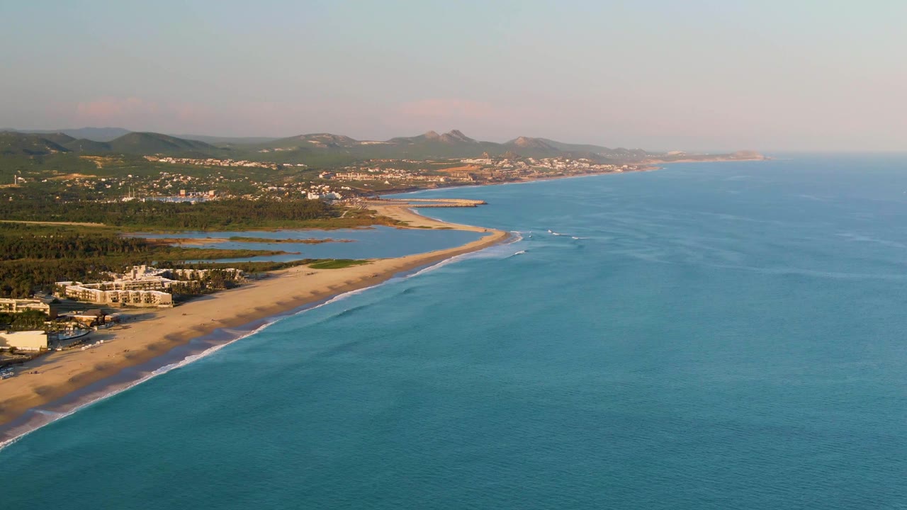 Aerial shoreline with turquoise water, sand, surf, buildings, lagoons, hills, and distant coast.