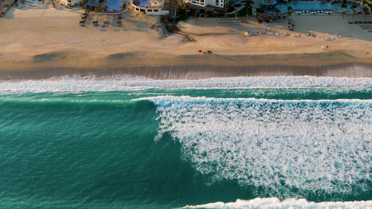 Aerial view in San Jose Del Cabo, Baja California Sur, with turquoise water and white foam moving toward a sandy beach, resort structures cropped along the top, and no visible people.