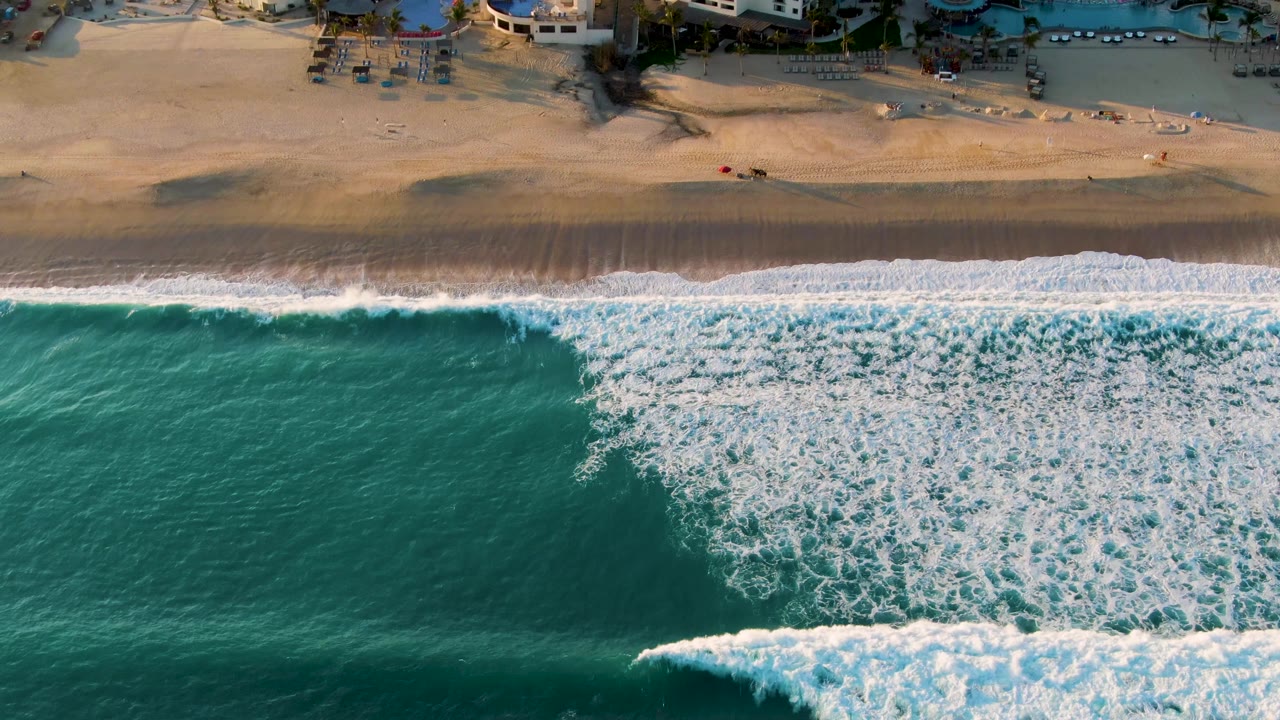 Aerial frame from San Jose Del Cabo with the breaking wave shifted farther right, deep green-blue water to the left, sandy beach above, and no visible people.