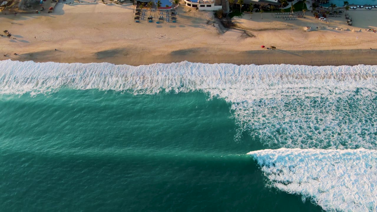 Overhead view in San Jose Del Cabo of turquoise ocean and a broad band of white foam approaching pale sand, with tiny beach figures and structures near the top edge.