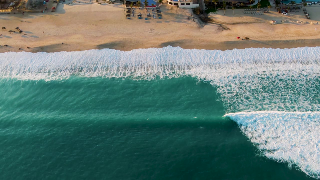 Aerial San Jose Del Cabo beach frame with a diagonal sheet of white foam crossing teal water and resort-edge buildings, chairs, and tiny people along the sand.