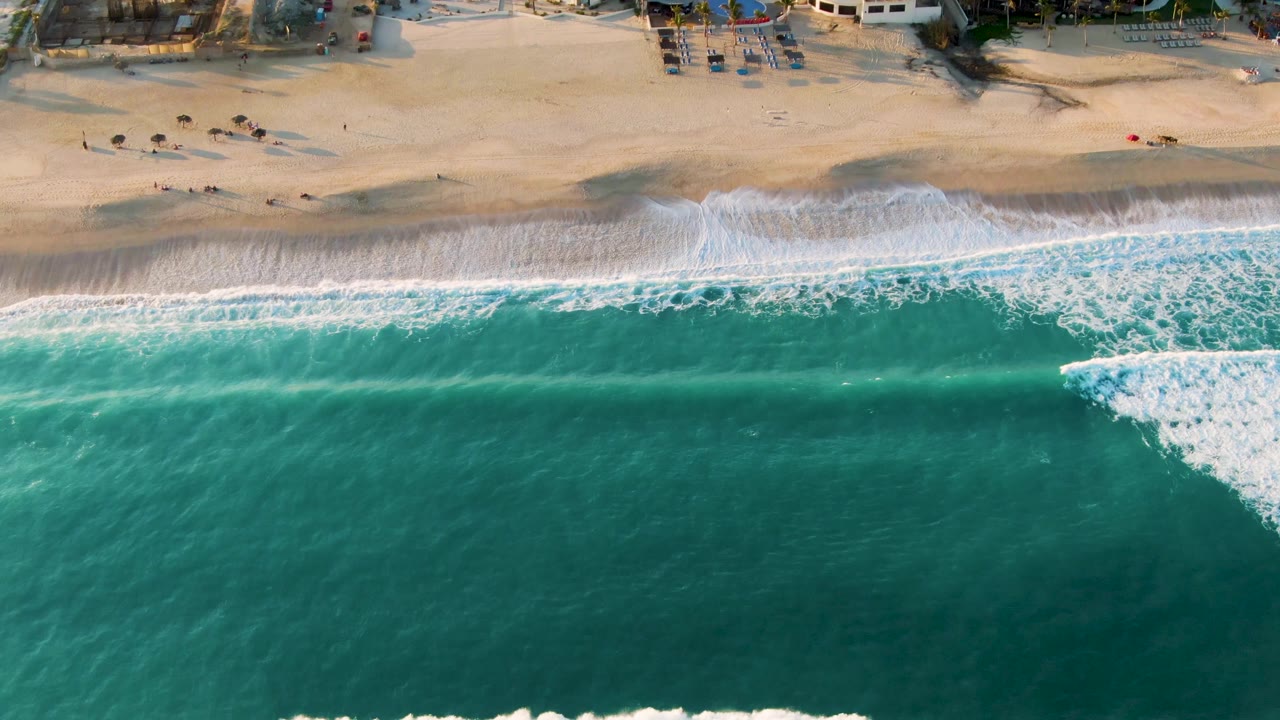 Overhead view in San Jose del Cabo, Mexico, with turquoise water filling the lower frame, a white breaking wave near the shoreline, tan sand above it, and small distant beachgoers near the top edge.
