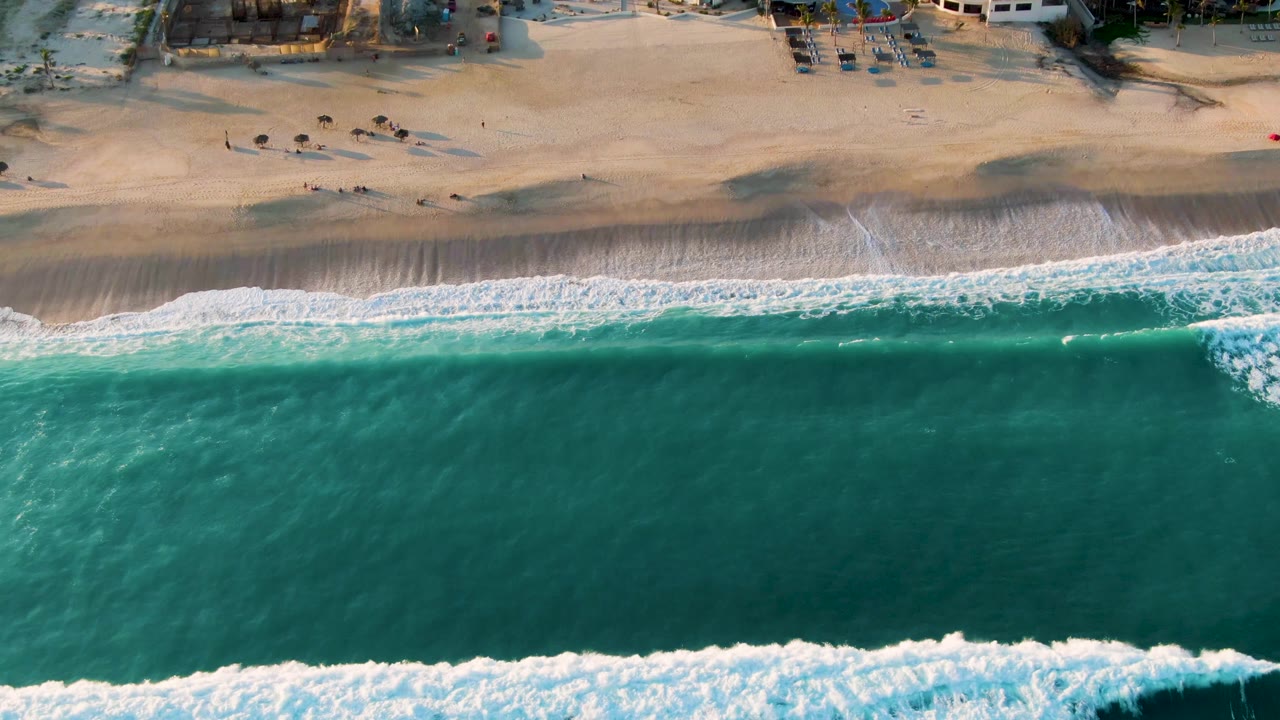 Top-down San Jose del Cabo shoreline view with a long white surf line, deep turquoise water below, pale sand above, and scattered tiny beachgoers and umbrellas near the top of frame.