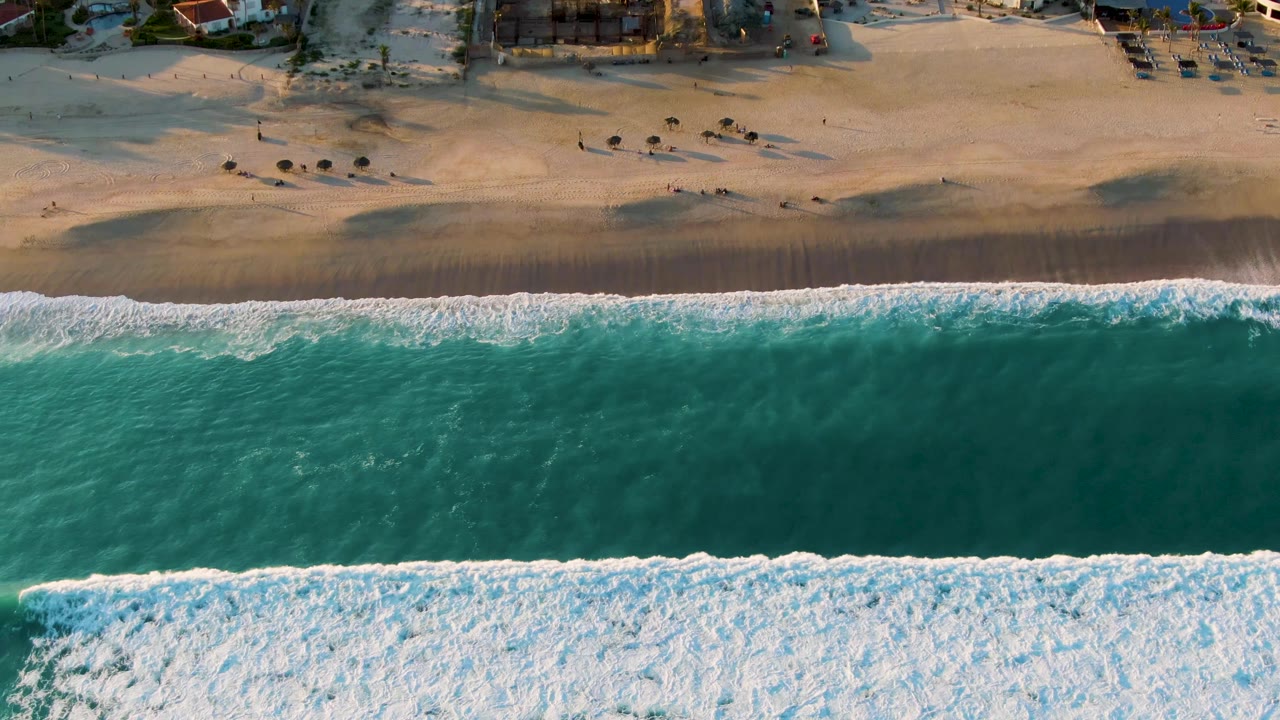 Overhead San Jose del Cabo beach view with turquoise water, a wide white foam band, pale sand, small beach umbrellas, beachfront buildings, and tiny visible people near the upper shore.