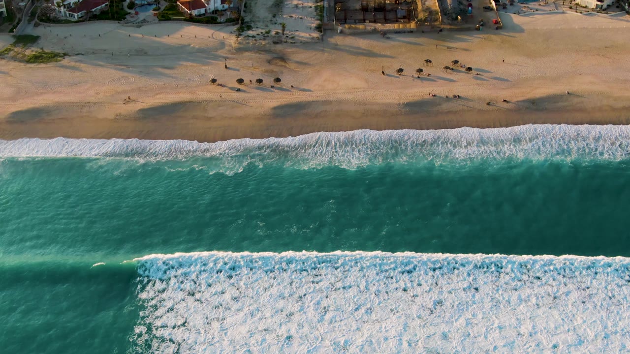 Top-down view of the San Jose del Cabo coast with foamy surf across the lower portion, green-blue water, light sand, scattered umbrellas, tiny beachgoers, and buildings along the top edge.