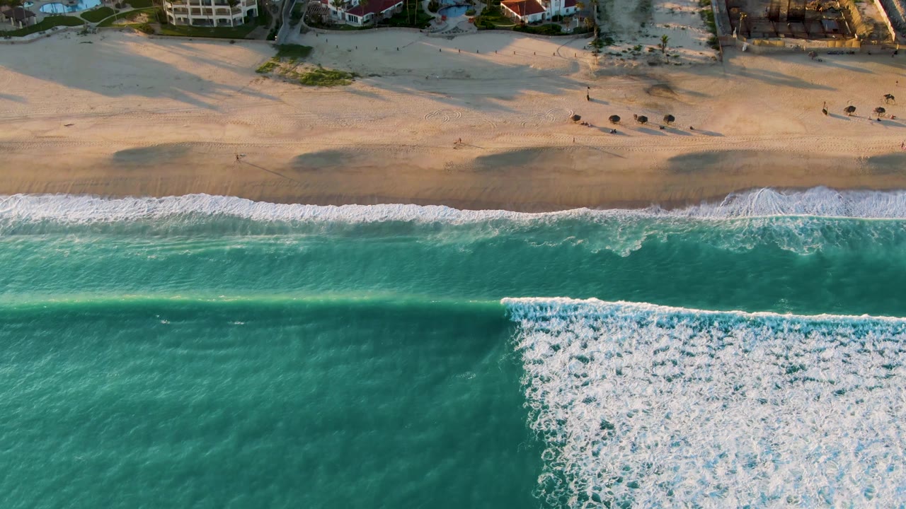 Overhead view of turquoise waves and white foam reaching a sandy San Jose Del Cabo beach, with beachfront houses along the top edge and no visible people.