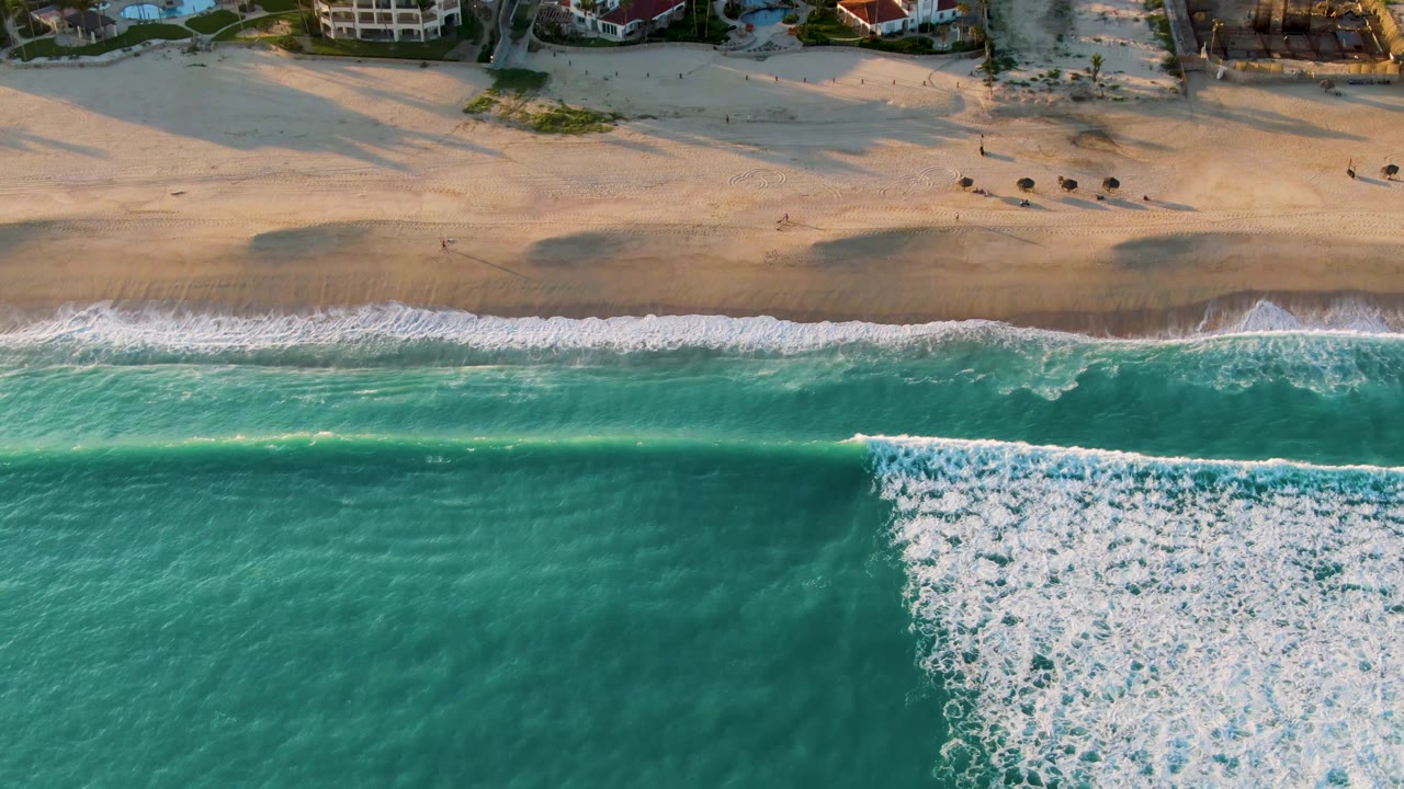 Aerial San Jose Del Cabo shoreline frame with a wider white foam patch spreading through turquoise water below the sandy beach, no visible people.