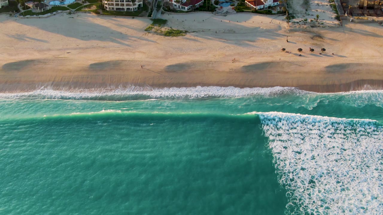 Top-down San Jose Del Cabo beach view where a thick white surf band sits near the right side of the turquoise water, with pale sand and beachfront homes above, no visible people.