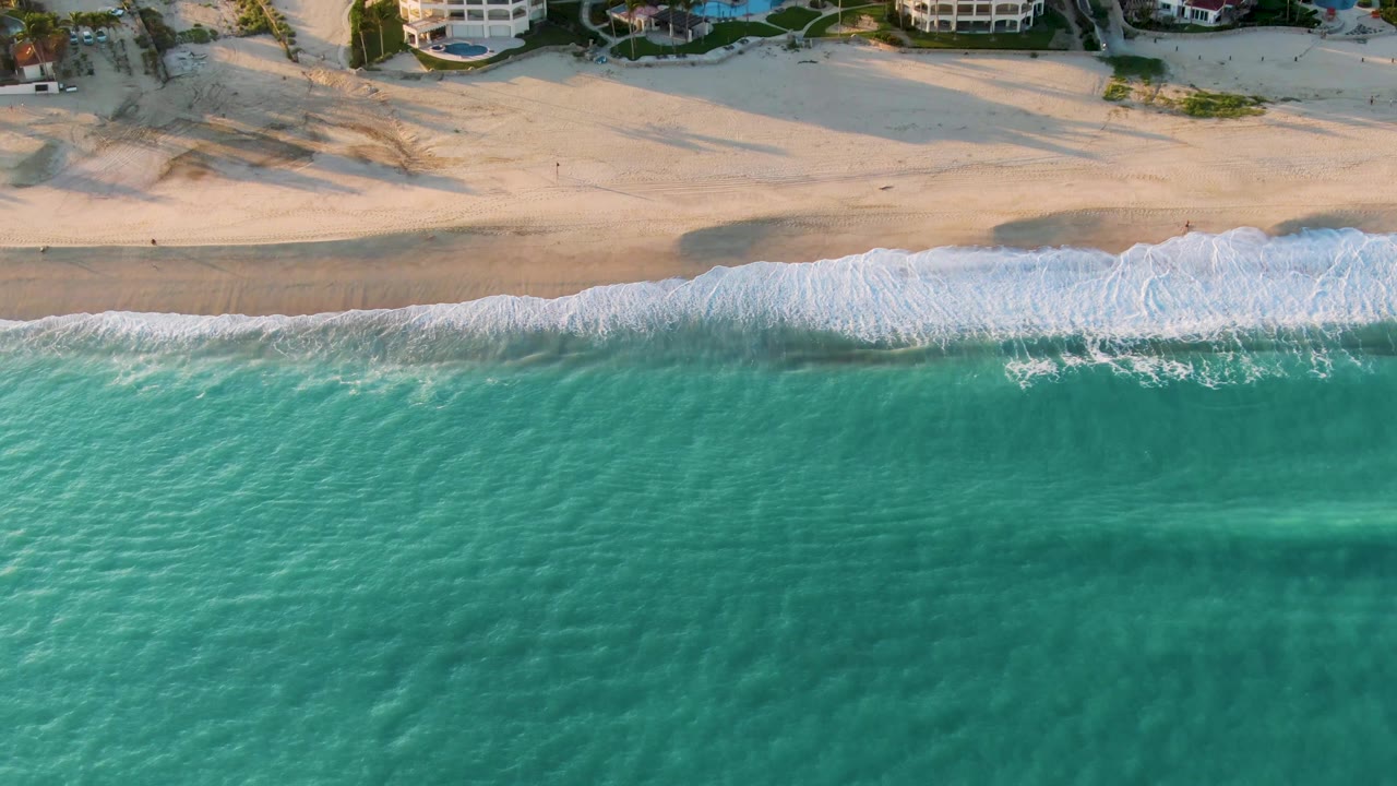 Overhead view in San Jose del Cabo, Mexico, with turquoise water below, a white surf line on wet sand, dunes, and beachfront buildings along the top edge, with no visible people.