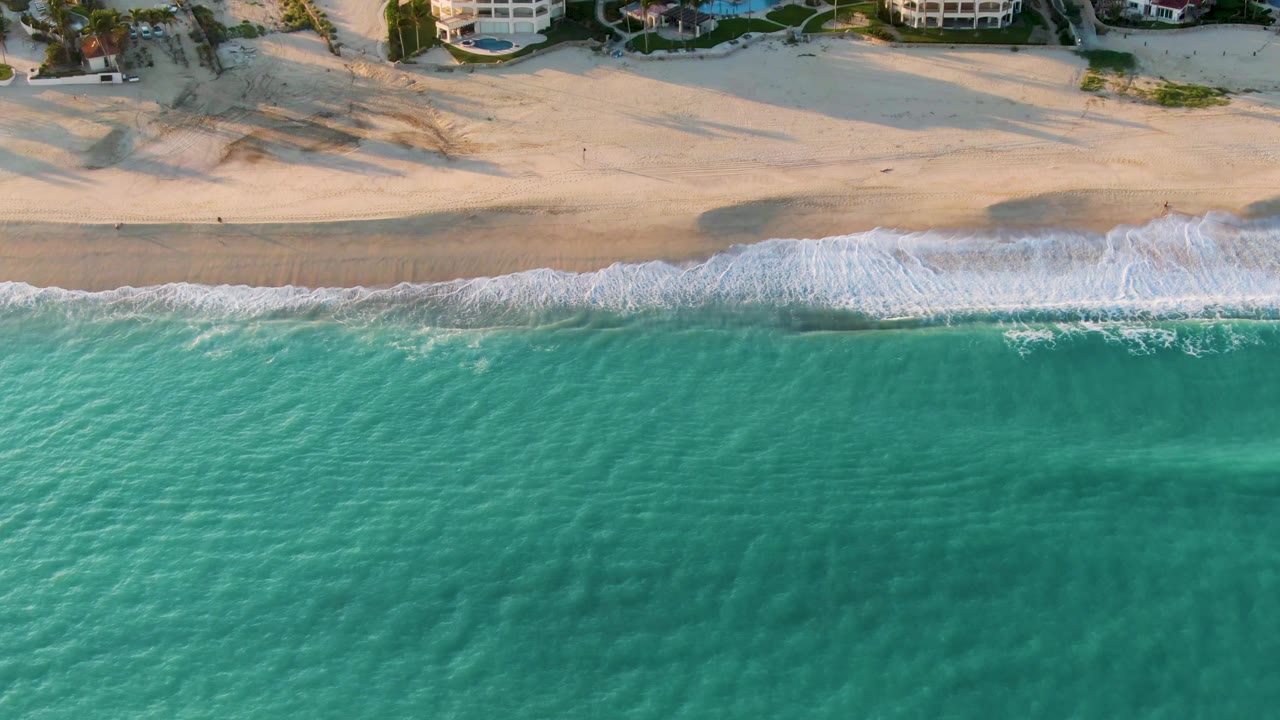 Aerial San Jose del Cabo beach frame showing a broader band of white foam curling onto wet sand, with dry dunes and beachfront buildings at the top and no visible people.