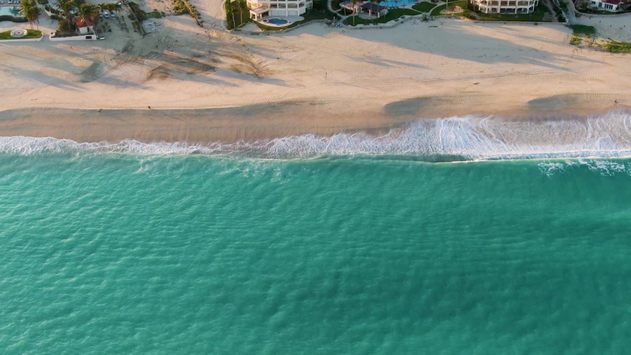 Overhead San Jose del Cabo shoreline view with the wave foam thinning into streaks over wet sand, teal water below, dunes above, and no visible people.
