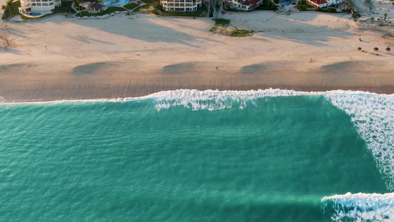 Aerial view over San Jose del Cabo turquoise water where a diagonal line of white surf reaches the sandy beach, with beachfront buildings at the top edge and no visible people.