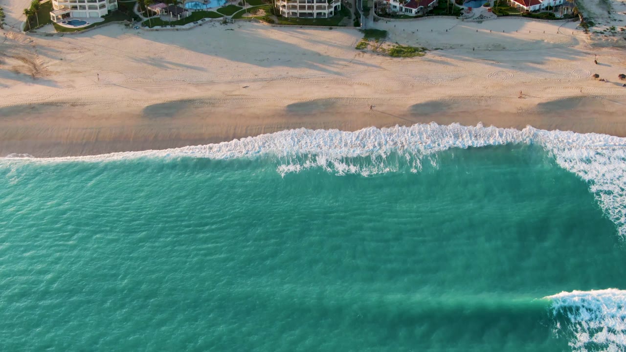 Aerial frame from San Jose del Cabo showing white wave foam advancing across the shoreline, turquoise water below, pale sand above, and no visible people.