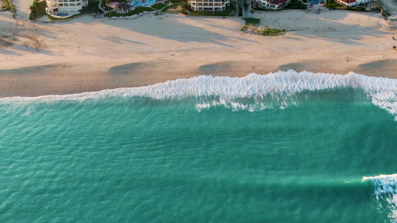 Aerial view in San Jose del Cabo with a broad turquoise water field, a broken white wave near the shore, sandy beach behind it, and no visible people.