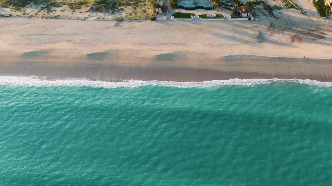 Overhead view in San Jose del Cabo showing turquoise water, a thin white surf line, pale sand, dune vegetation, and no visible people at the start of the clip.