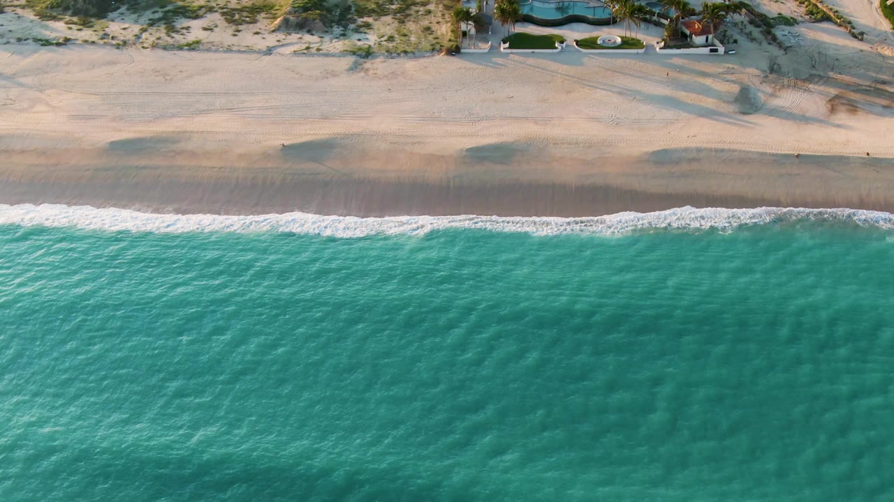 San Jose del Cabo aerial frame with clear turquoise water below, a narrow band of foam on the shore, pale beach above it, and no visible people.