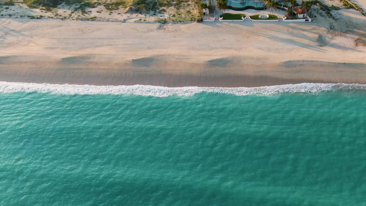 Overhead San Jose del Cabo coastline with rippled turquoise water, soft surf, a broad empty beach, dune vegetation, visible pool areas, and no visible people.
