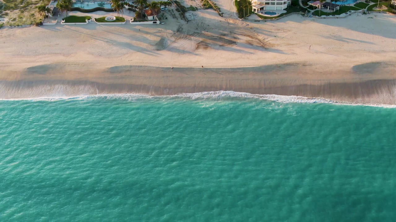 Aerial frame over San Jose Del Cabo showing turquoise water, a thin surf line, pale sand, dune vegetation, beachfront homes, and no visible people.