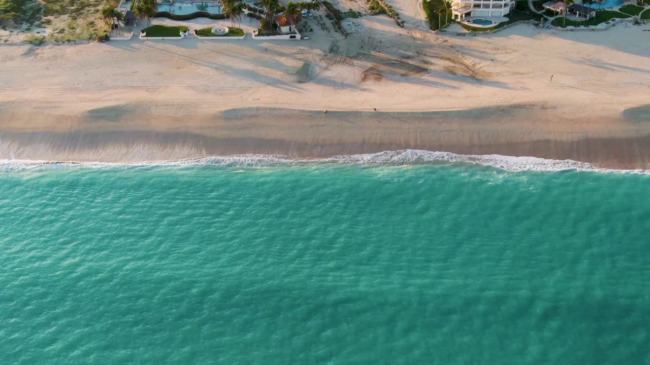 Aerial San Jose Del Cabo shoreline frame with rippled turquoise water, white foam on the wet sand, dune shadows, coastal vegetation, houses, and no visible people.