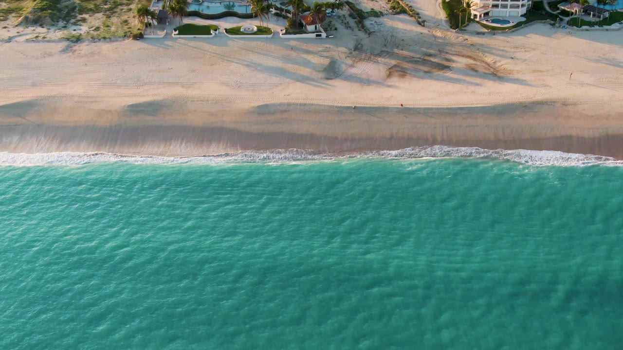 Aerial frame from San Jose Del Cabo with the ocean occupying most of the view, a narrow white wave edge, broad empty sand, dunes, houses, and no visible people.