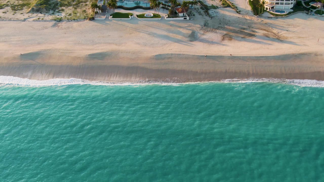 Beachfront houses and dune vegetation sit above a sandy shore and turquoise water.