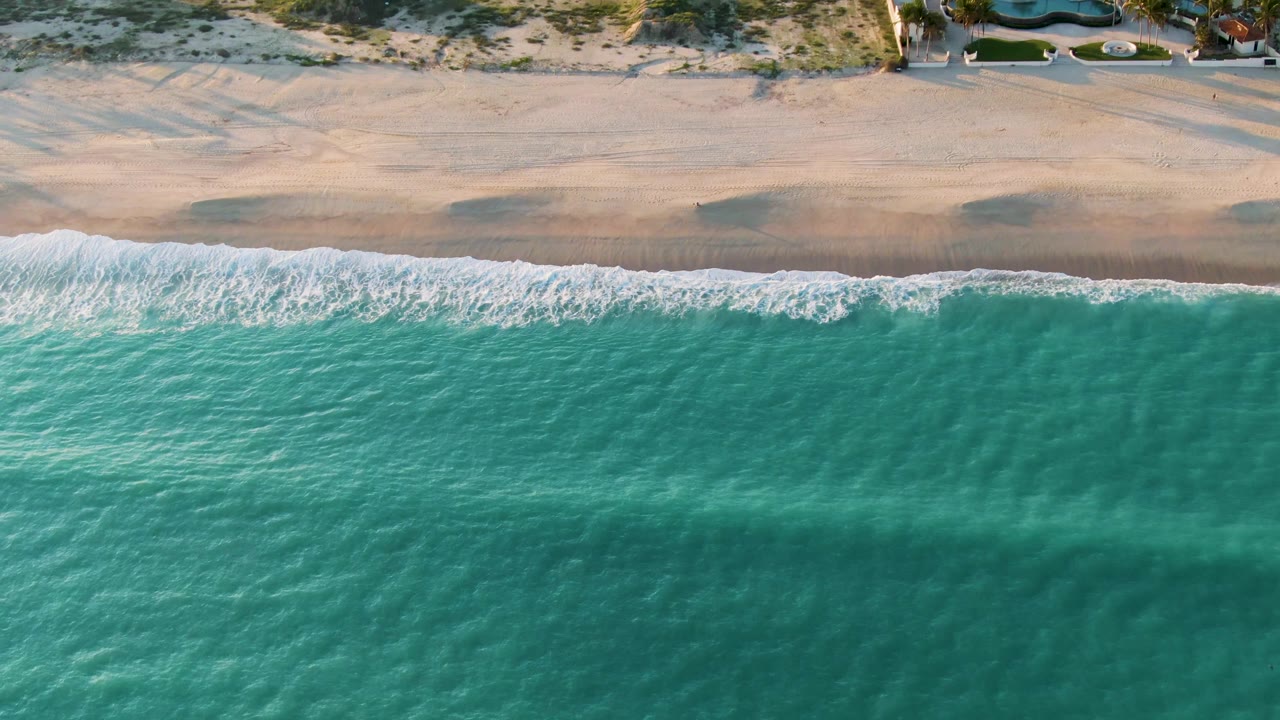 Overhead view in San Jose Del Cabo, Mexico, with turquoise water pushing white foam onto a pale sandy shore and sparse dunes beyond, with no visible people.