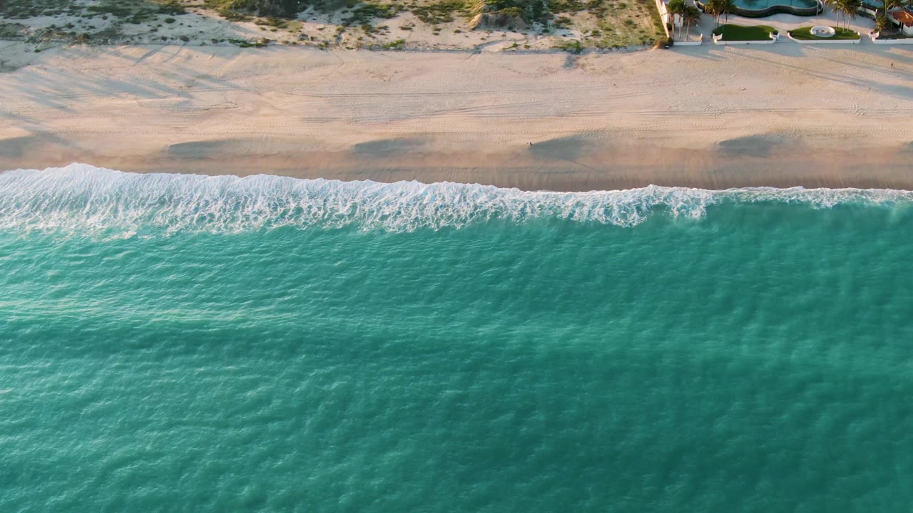 Aerial frame from San Jose Del Cabo, Mexico, showing a broad band of turquoise water, a thin white surf line, pale beach sand, and dunes with no visible people.