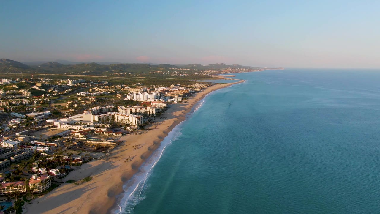 Aerial view over San Jose Del Cabo with a broad sandy beach on the left, blue ocean on the right, coastal buildings near shore, inland hills, and no clearly visible people at this moment.
