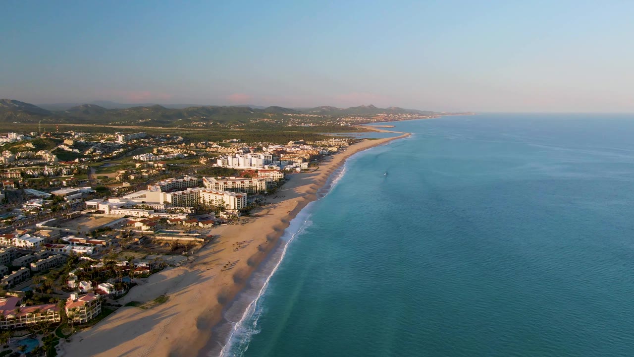Wide aerial coastline with beach, town buildings, ocean, and a faint offshore mark.