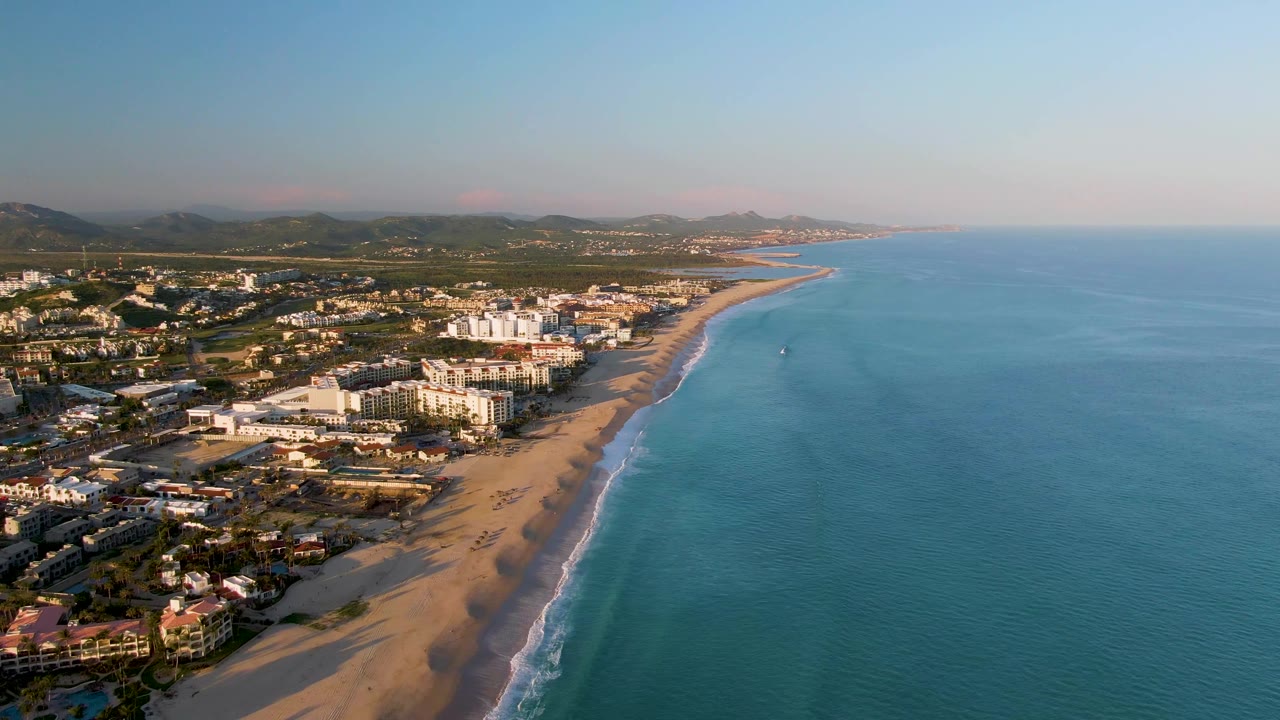 Aerial view of San Jose Del Cabo coastline with warm light on beach buildings, a long strip of sand, gentle surf, blue water, and a small distant person or water user offshore.