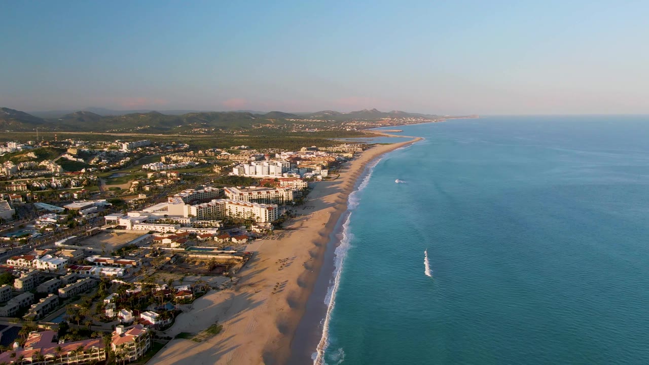 Aerial view over San Jose Del Cabo with sandy beach, coastal buildings, blue ocean, distant hills, and no visible people at the start of the clip.