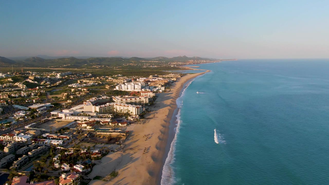 Wide aerial view of San Jose Del Cabo coastline with clustered buildings, a broad sandy beach, calm blue water, low hills, and no visible people.