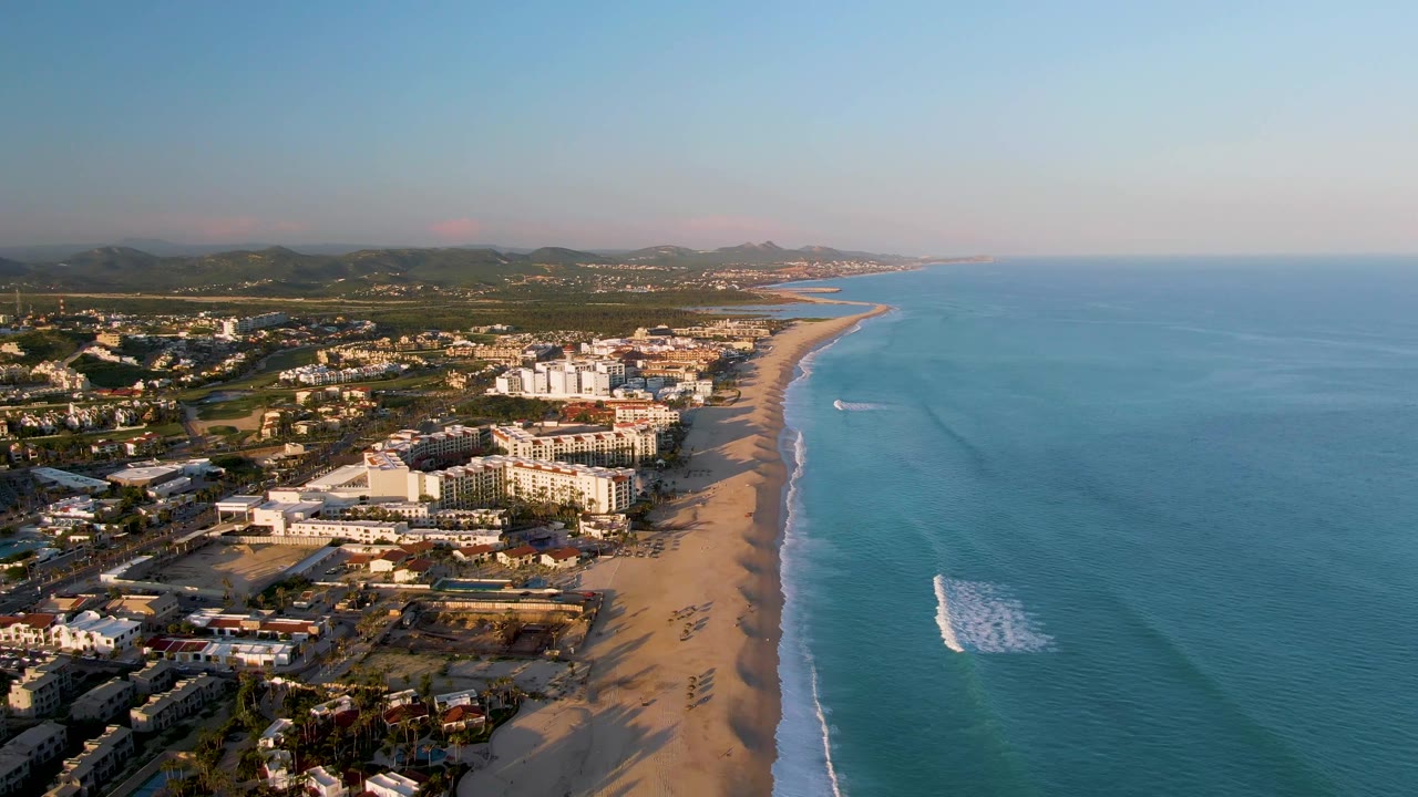 Aerial view over San Jose del Cabo with blue ocean on the right, a sandy beach below, low buildings along the shore, distant hills, and no visible people.