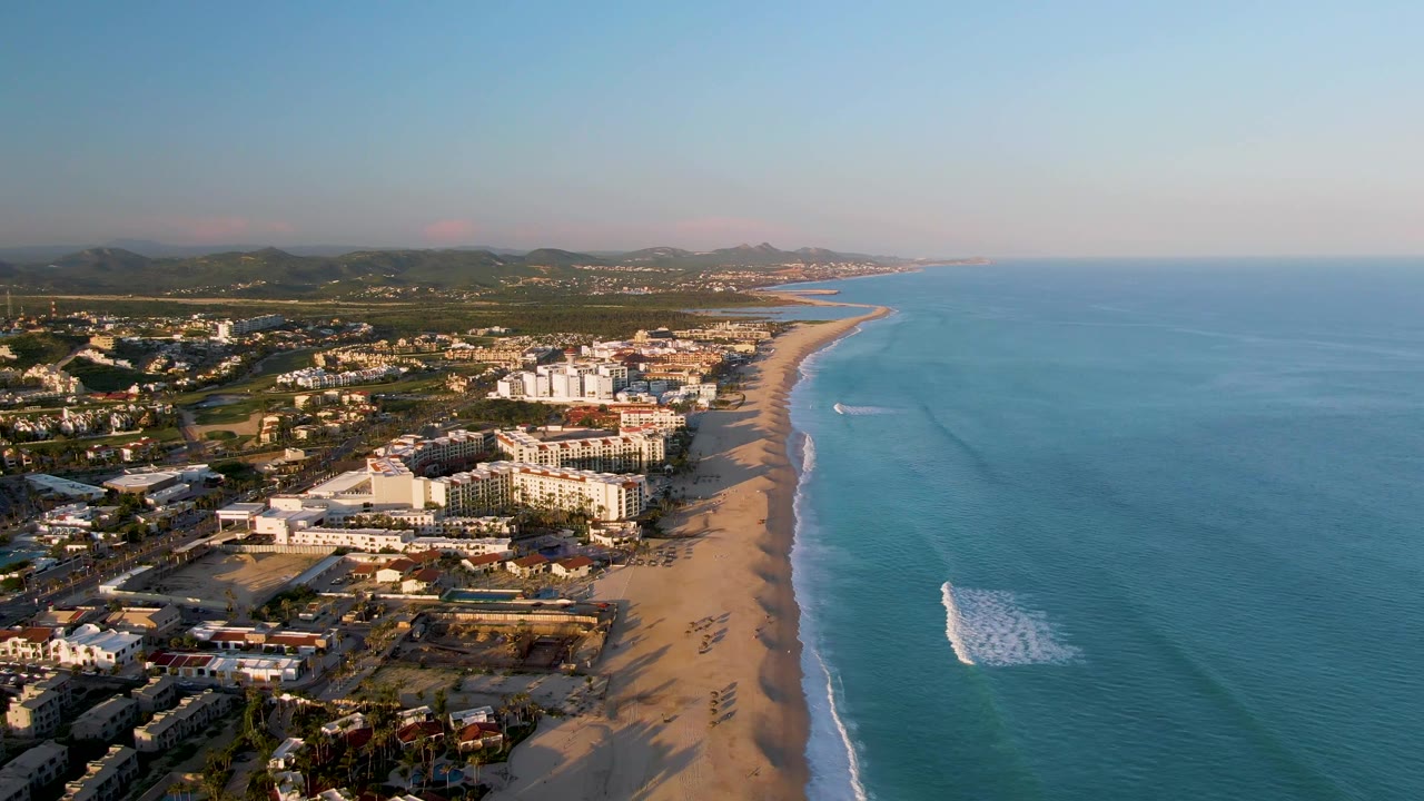 Wide aerial coastline with beach surf and buildings beside the shore.