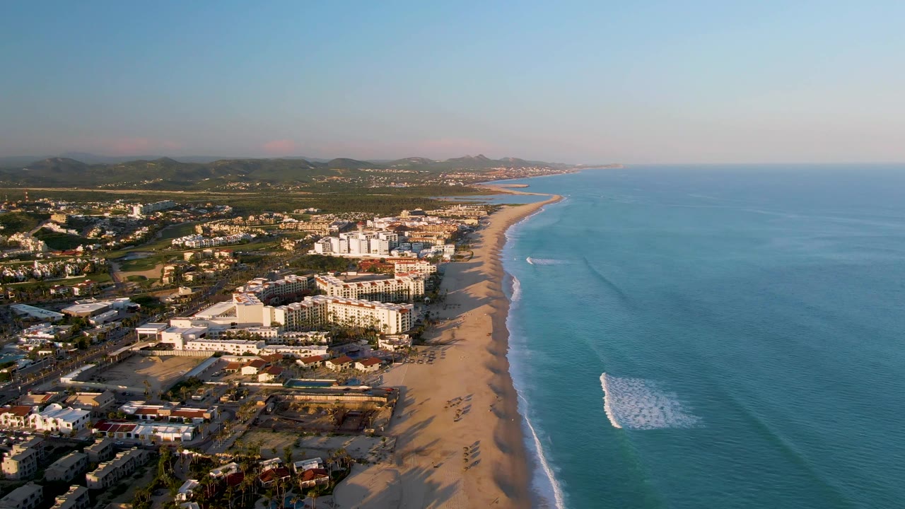 Drone-style view of San Jose del Cabo beach with turquoise-blue water, a narrow surf band, shorefront buildings, inland roads, low hills in the background, and no visible people.