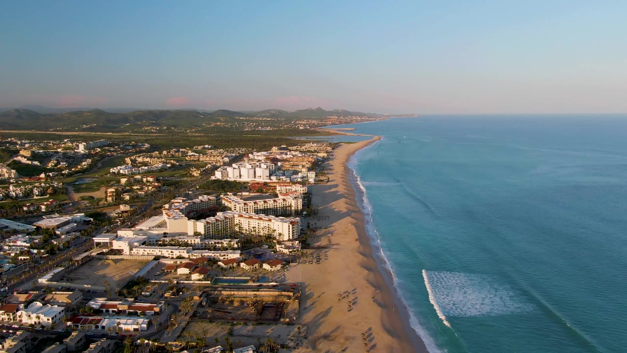Aerial frame over San Jose Del Cabo showing beachfront buildings beside a long sandy beach, blue ocean, white surf, inland hills, and no visible people.