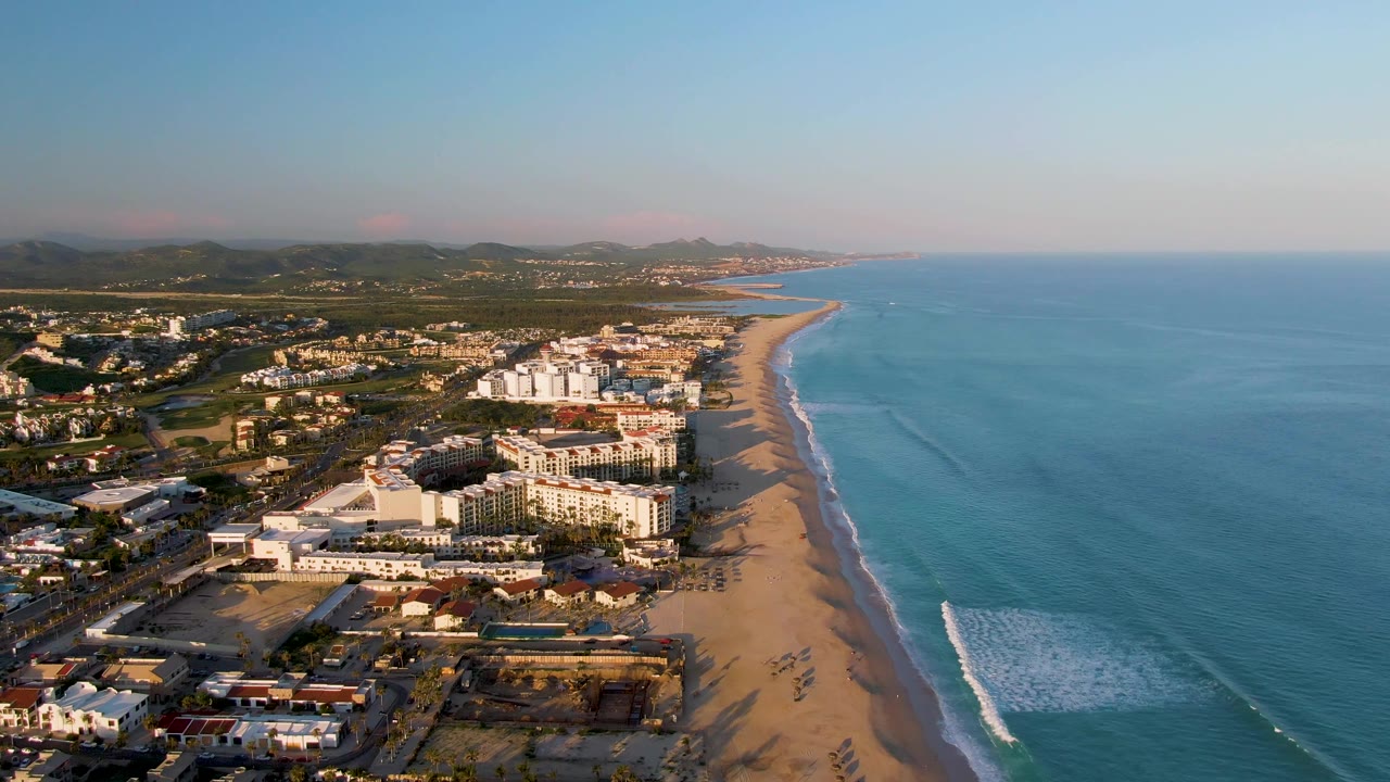 High aerial frame in San Jose Del Cabo with the waterfront buildings slightly more centered, the beach curving into the distance, blue water to the right, and no visible people.