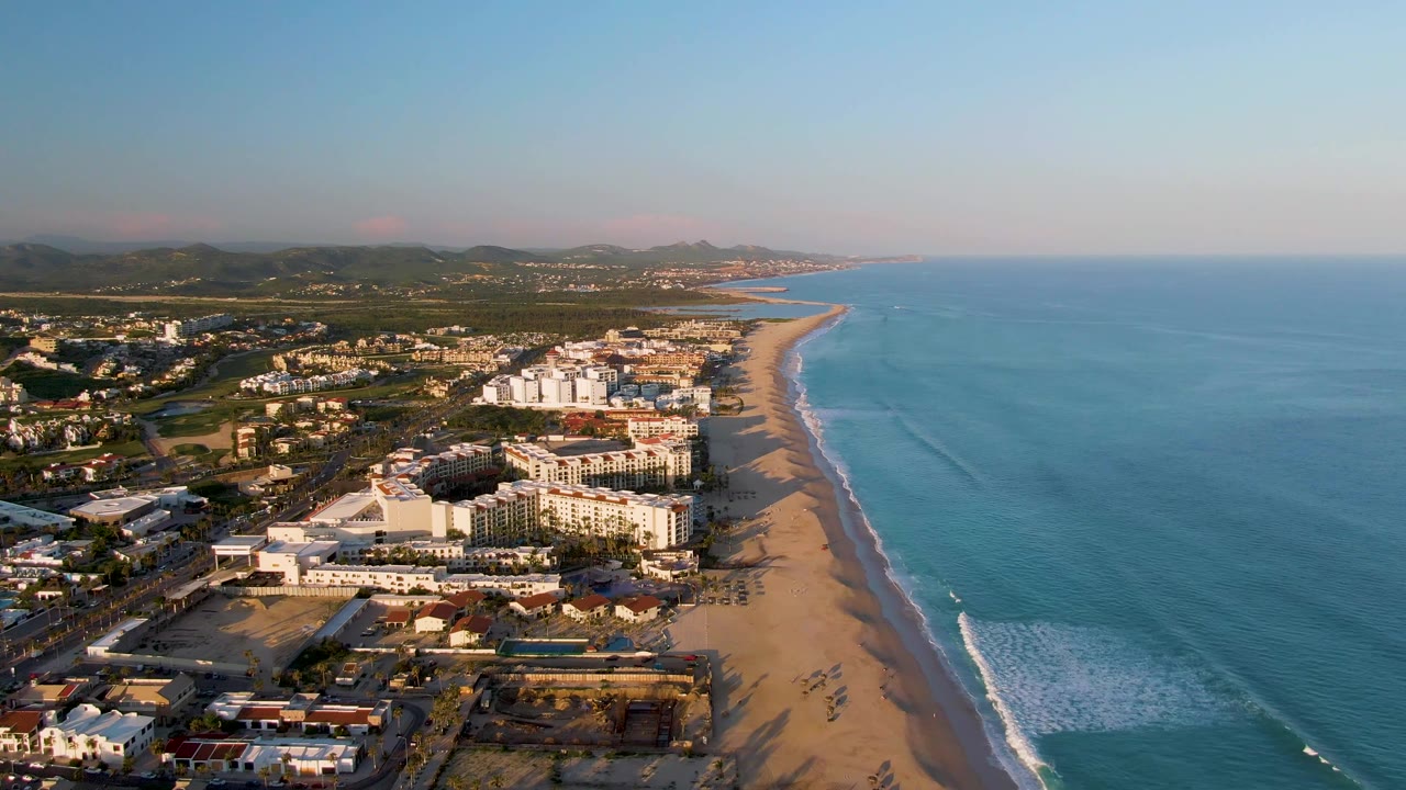 Aerial view over San Jose Del Cabo with clustered beachfront buildings in warm light, a broad strip of sand, small breaking waves, distant hills, and no visible people.
