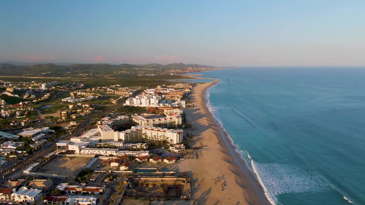 Wide aerial coastline with buildings, beach, surf, and inland hills