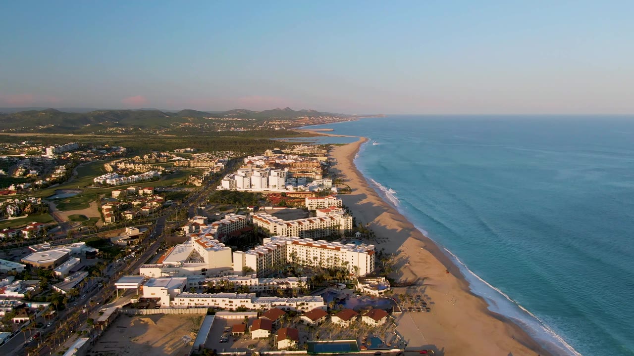 Aerial dawn view over San Jose del Cabo resort buildings, sandy beach, blue ocean, distant hills, and no visible people.
