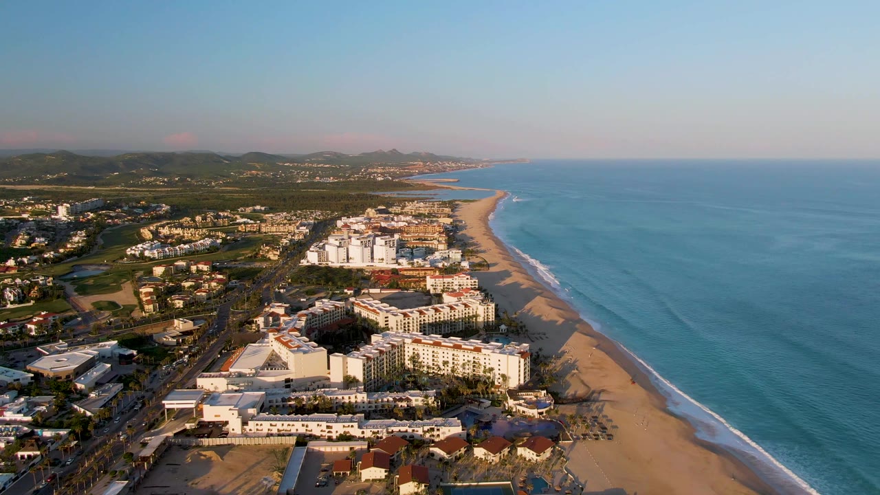 Wide aerial frame of San Jose del Cabo coastline with white resort buildings, tan sand, blue water, soft dawn light, and no visible people.