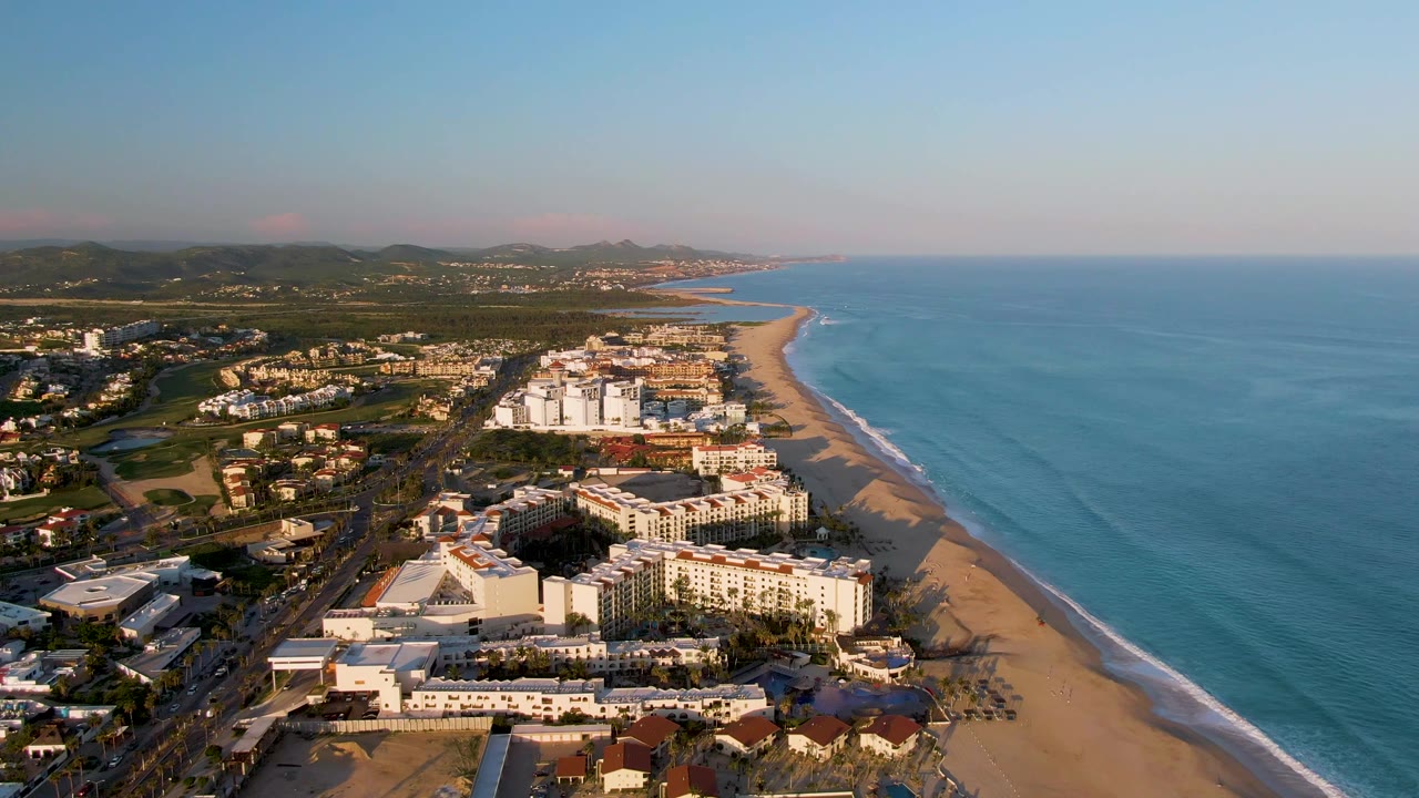 Drone-like view over San Jose del Cabo beachfront hotels with ocean on the right, roads and hills on the left, and no visible people.