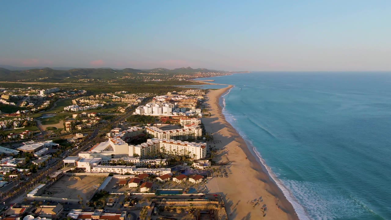 Aerial coastline view with beachfront resort buildings, sand beach, blue ocean, and inland hills.