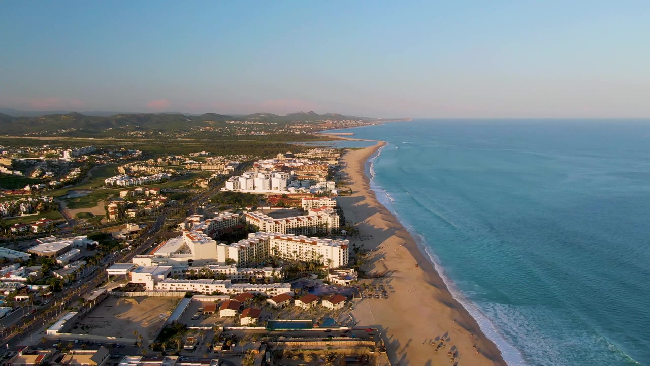 Drone view over San Jose Del Cabo beachfront resort blocks, with the shoreline running into the distance, ocean water on the right, mountains on the horizon, and no visible people.