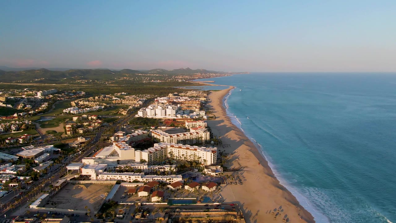 Wide aerial frame from San Jose Del Cabo showing ocean occupying the right half, a long beach and resort frontage through the center, low mountains beyond town, and no visible people.