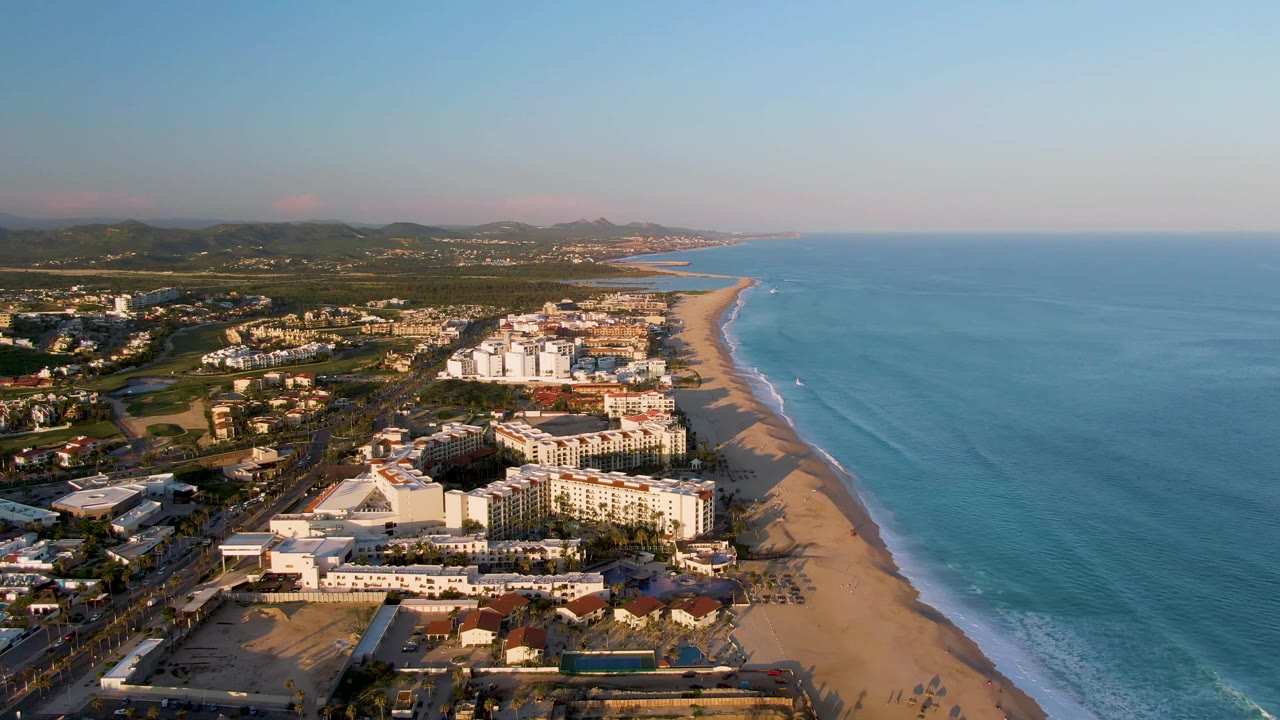 Aerial view along the San Jose Del Cabo resort beachfront, with angled warm light on white buildings, surf lines on the sand, inland hills to the left, and no visible people.