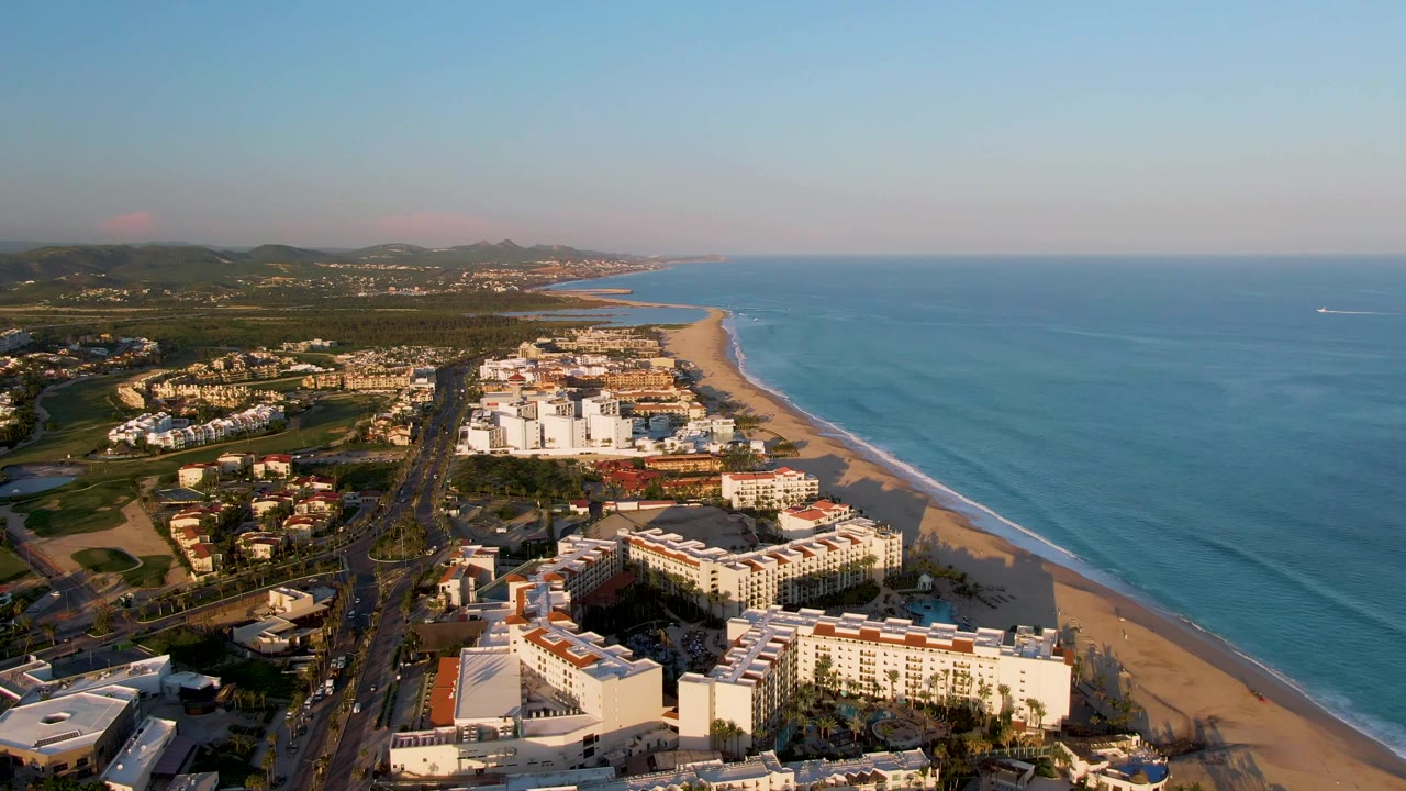 Aerial frame over San Jose Del Cabo showing white beachfront resort buildings, a coastal road, sandy beach, blue ocean, distant hills, and no visible people.