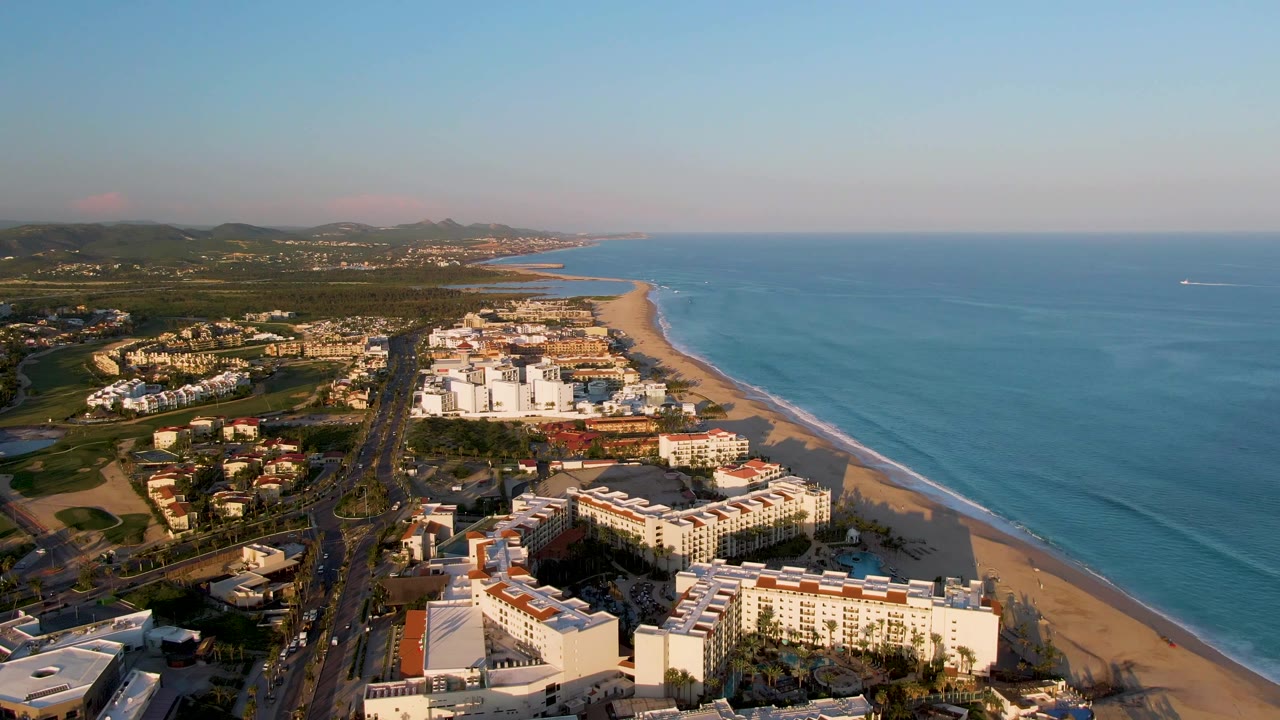 A high aerial view in San Jose Del Cabo shows the resort blocks and coastal road slightly farther along the shoreline, with ocean filling the right side and no visible people.